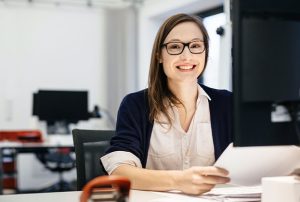 A young businesswoman wearing glasses is sitting in front of a computer in an office room, holding some paper while smiling happily.
