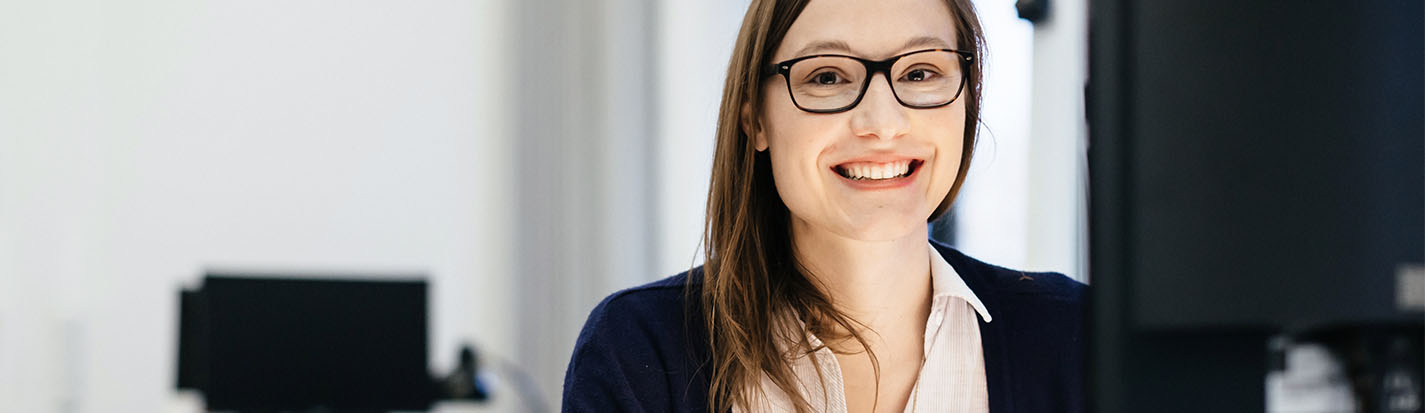 A young businesswoman wearing glasses is sitting in front of a computer in an office room, holding some paper while smiling happily.