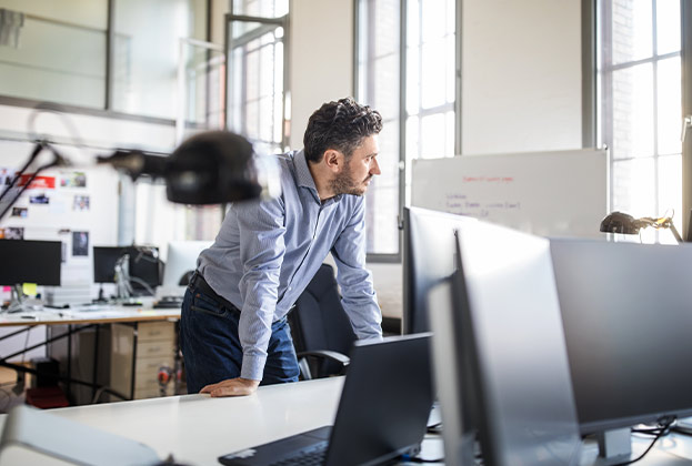 Thoughtful businessman leaning on desk by computer monitor while looking away at creative office