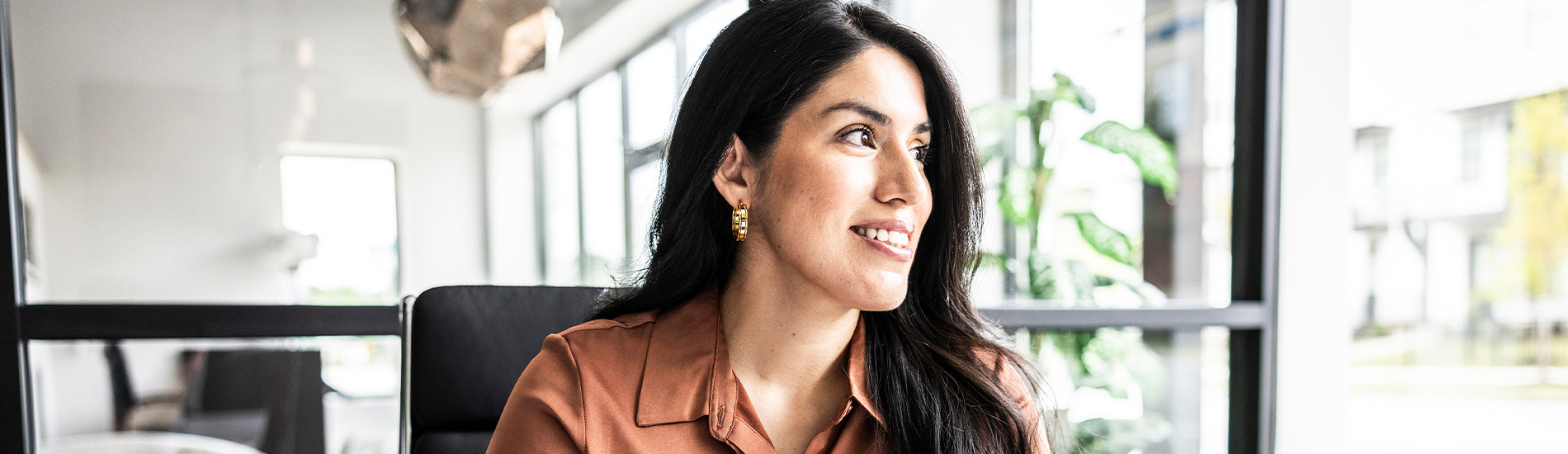 A woman wearing an orange shirt, sitting at a desk and looking out a window.