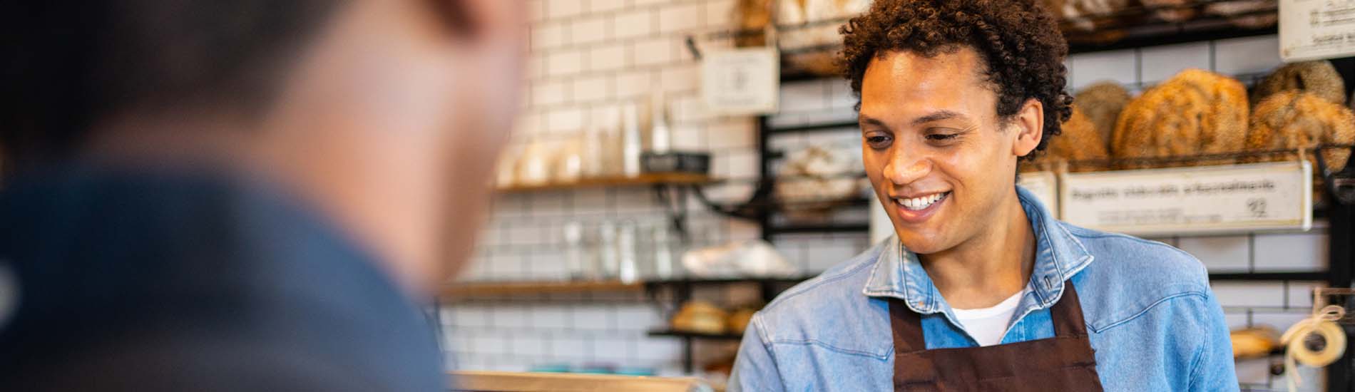 A friendly baker, wearing an apron, interacts with a customer, handing them a bag of bread in a warm and inviting bakery setting.
